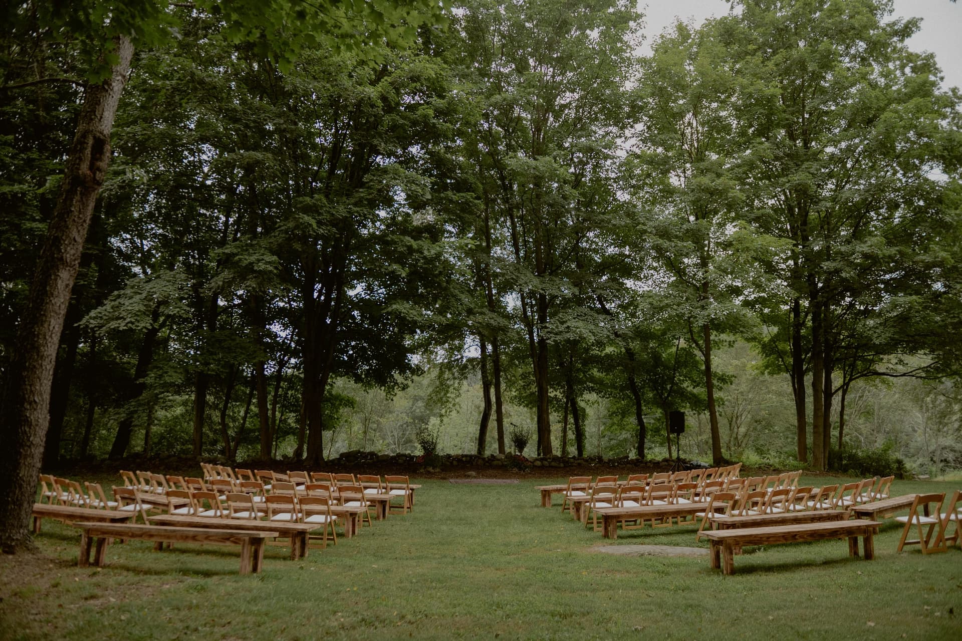 Outdoor wedding ceremony setup with wooden benches arranged in a semicircle surrounded by tall trees and lush greenery, photo by Chellise Michael Photography
