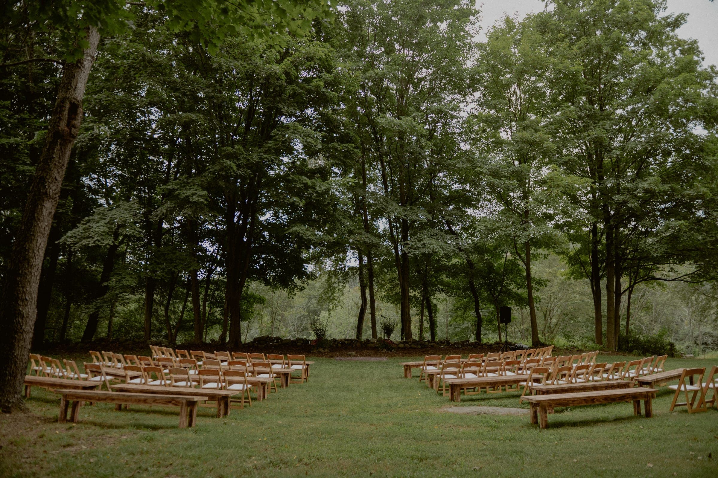Outdoor wedding ceremony with wooden benches and chairs arranged in a semicircle on a grass lawn surrounded by mature trees, photo by Chellise Michael Photography